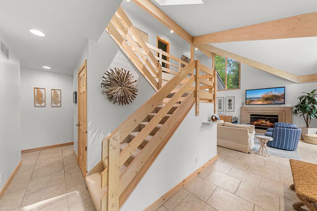 A bright foyer area with light tiled flooring and a prominent wooden staircase with open risers, leading to an upper level, with a view into a spacious living room featuring a fireplace and large windows.