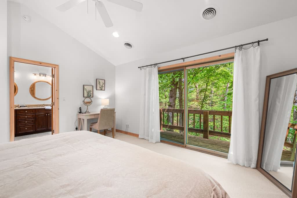 Bright bedroom with a king-sized bed, light carpet, and large sliding glass doors opening to a private deck with views of surrounding trees. A full-length mirror stands against the wall.