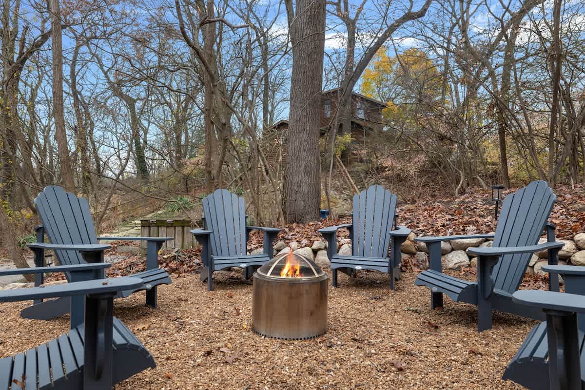 Outdoor fire pit area with a circular arrangement of five gray Adirondack chairs on a gravel ground, surrounded by bare trees and natural landscaping. A modern, cylindrical fire pit is at the center.