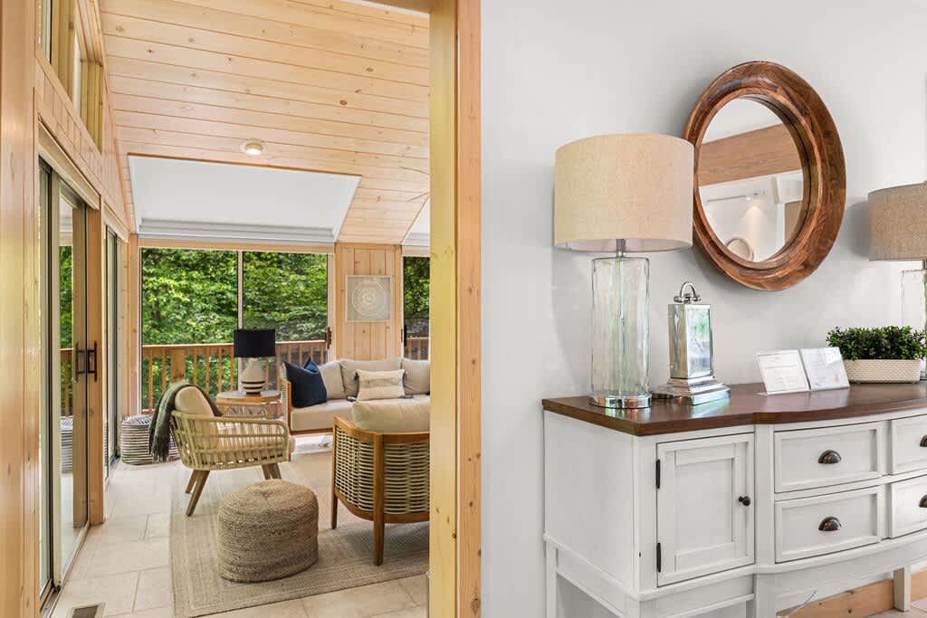 A bright hallway with light tiled flooring, featuring a white console table adorned with lamps and a round wooden mirror, leading into a sun-drenched screened porch with wooden walls and large windows.