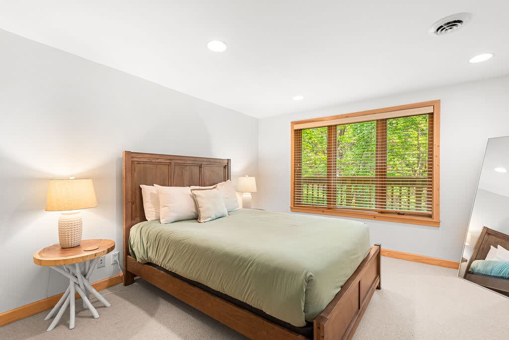 Bright bedroom featuring a wooden queen bed with a tall headboard and light green bedding, flanked by unique wooden nightstands with textured lamps. A large window with wooden blinds and trim overlooks a vibrant green outdoor scene.