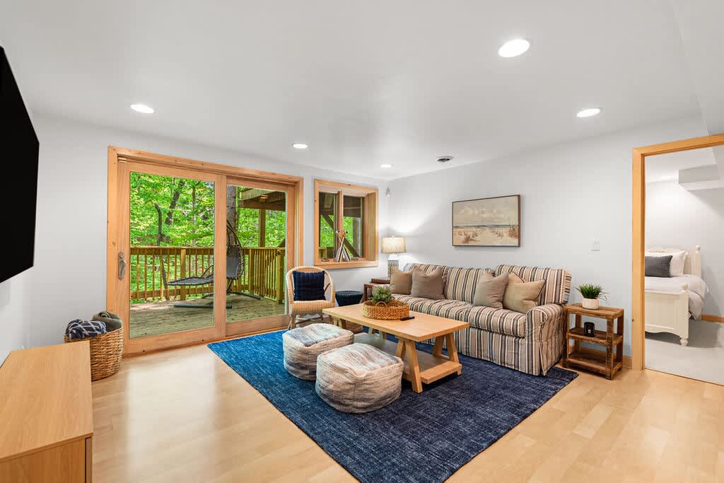 Cozy living space with a striped sofa, wooden coffee table, blue area rug, and sliding glass doors leading to a deck with lush green trees visible outside.