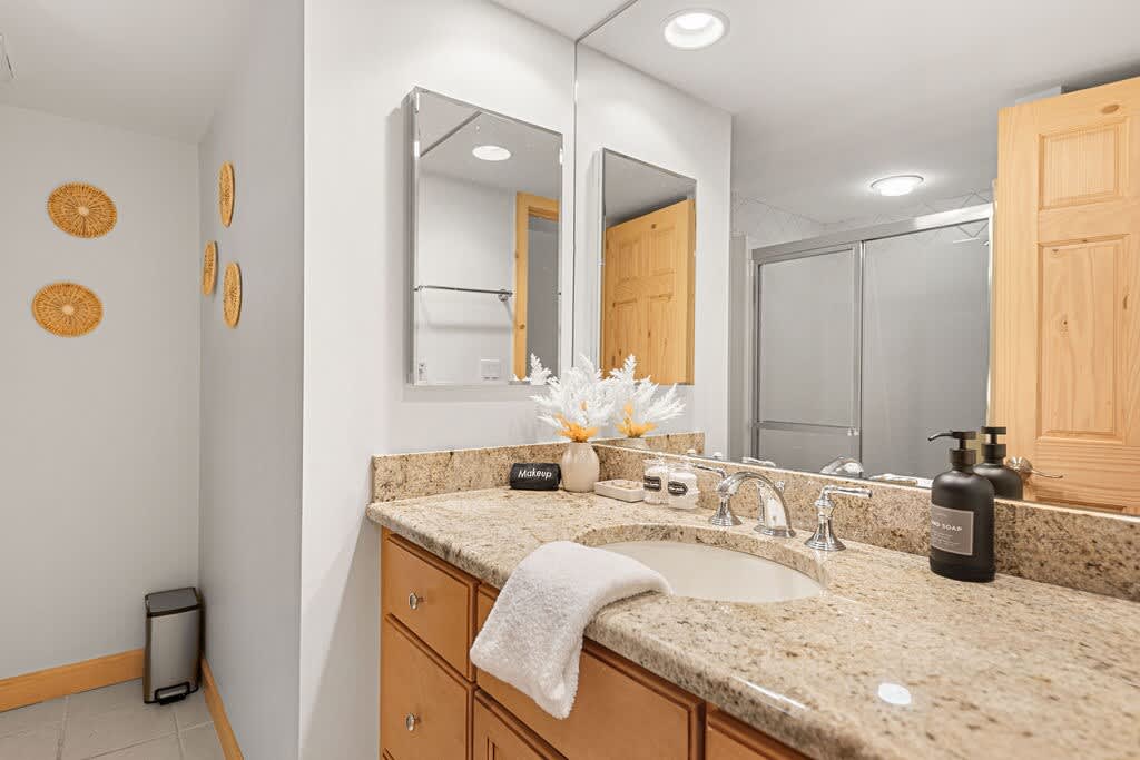 Modern bathroom featuring a light granite countertop with an undermount sink, light wood vanity, and two rectangular mirrors reflecting a shower and wooden door. Decorative woven wall hangings add a touch of texture to the white walls.