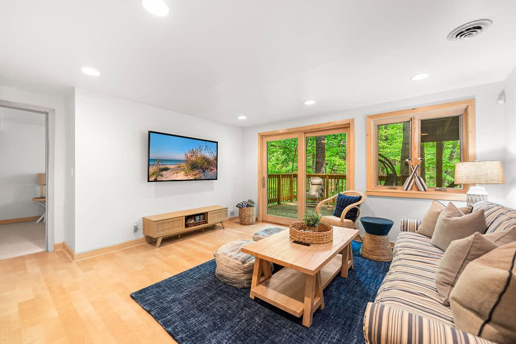 Bright living room featuring a striped sofa, wooden coffee table, blue area rug, and a large wall-mounted TV, with sliding glass doors and a window offering views of a wooded deck.