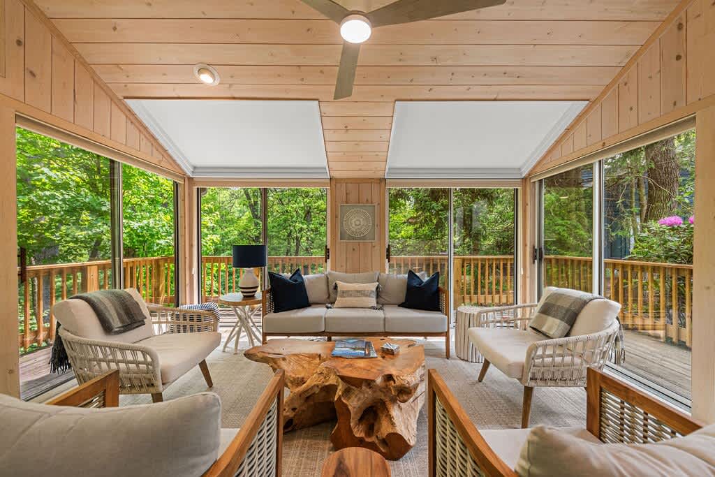 A cozy screened porch with light wooden paneling on walls and ceiling, furnished with comfortable seating, a unique tree-trunk coffee table, and surrounded by large windows offering panoramic views of lush green trees.
