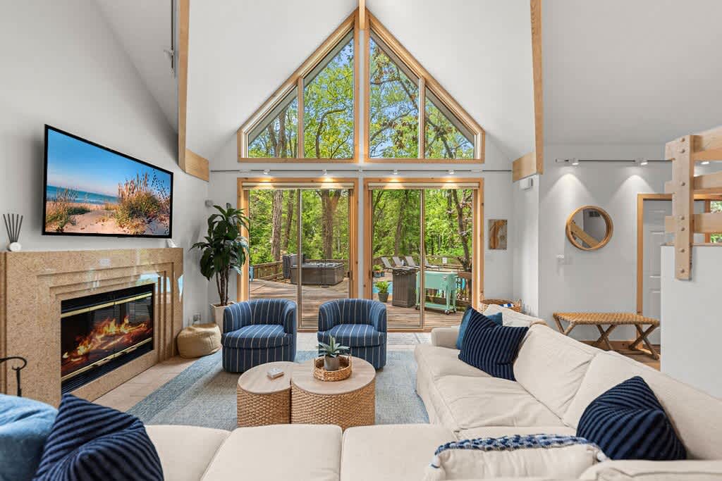 Spacious living room with a high vaulted ceiling and a wall of A-frame windows overlooking the lush outdoor greenery. A large sectional sofa and two blue striped armchairs are arranged around a stone fireplace with a mounted TV.