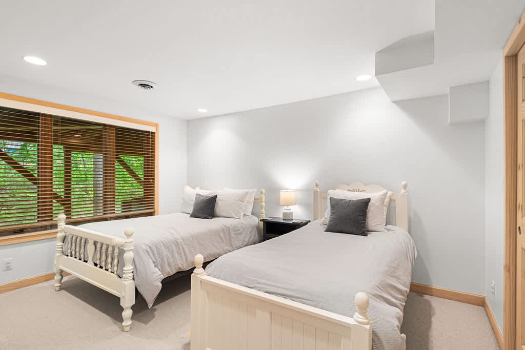 Cozy bedroom with two white twin beds featuring decorative headboards and footboards, dressed in light gray bedding. A large window with wooden blinds and trim provides natural light, and a small nightstand with a lamp sits between the beds.