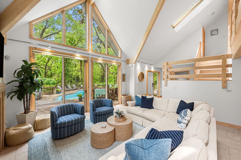 Bright living room featuring a vaulted ceiling with exposed wooden beams and a large A-frame window. A cream sectional sofa with blue accent pillows faces a stone fireplace, with a loft area visible above.