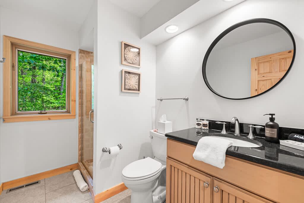 Contemporary bathroom with a dark granite countertop, undermount sink, and light wood vanity with slatted details. A large round mirror hangs above the sink, and a glass-enclosed shower is visible through a window with natural wood trim.