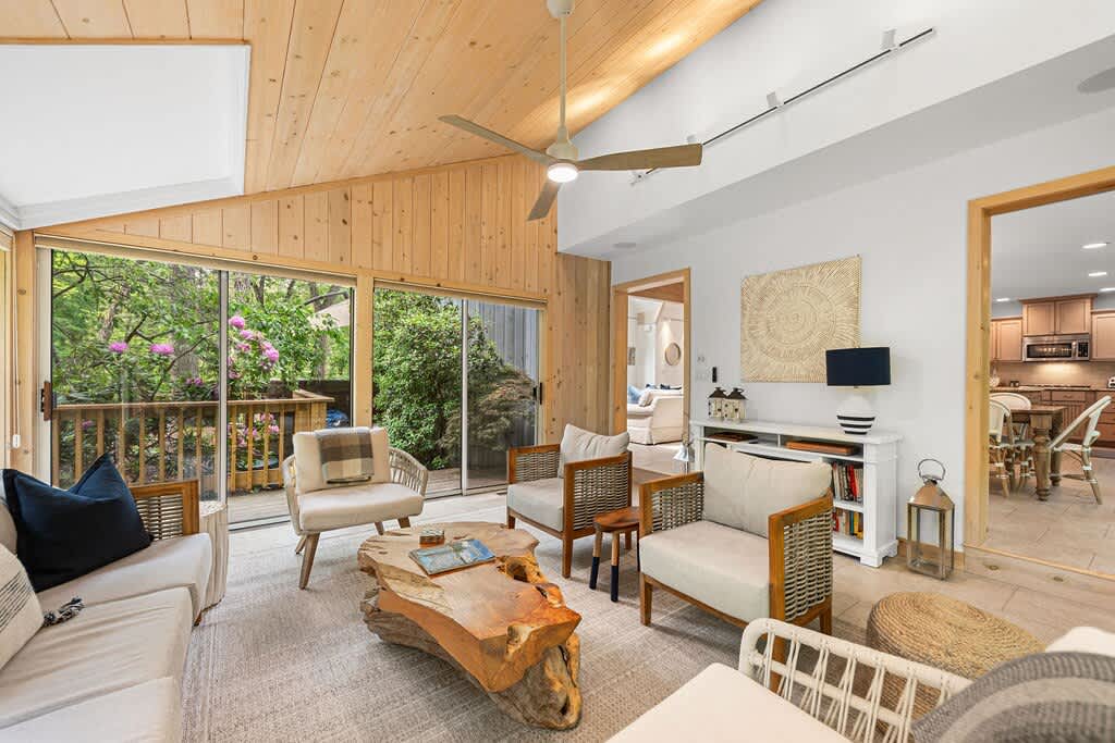 A spacious living room featuring a vaulted wooden ceiling, large windows that open to a lush outdoor view, and comfortable seating arranged around a distinctive tree-trunk coffee table, with a glimpse of the kitchen/dining area.