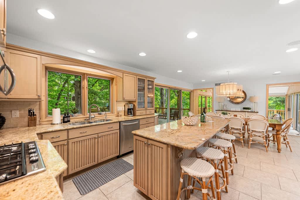 A spacious kitchen with light wood cabinetry and speckled granite countertops, featuring a large island with bar seating, a double sink under a window, and stainless steel appliances, all set on light tiled flooring.