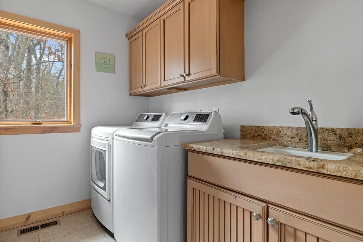 Functional laundry room equipped with a white top-loading washing machine and front-loading dryer, set beneath light wood upper cabinets. A granite countertop with an undermount sink and a window with natural wood trim complete the space.