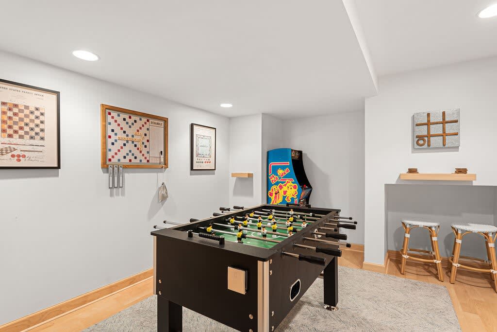 Recreation room featuring a black foosball table on a light rug, with light gray walls adorned with framed artwork. An arcade game machine and two small stools are also visible, suggesting a fun entertainment space.