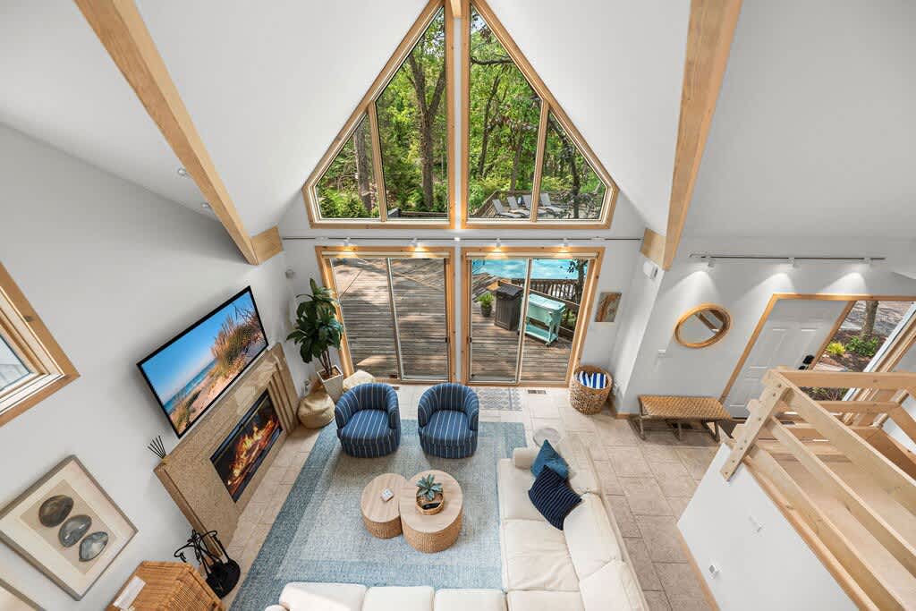 High-angle view of a bright living room with a vaulted ceiling, exposed wooden beams, a large A-frame window overlooking trees, a fireplace, and a mix of seating including a white sectional and blue armchairs.