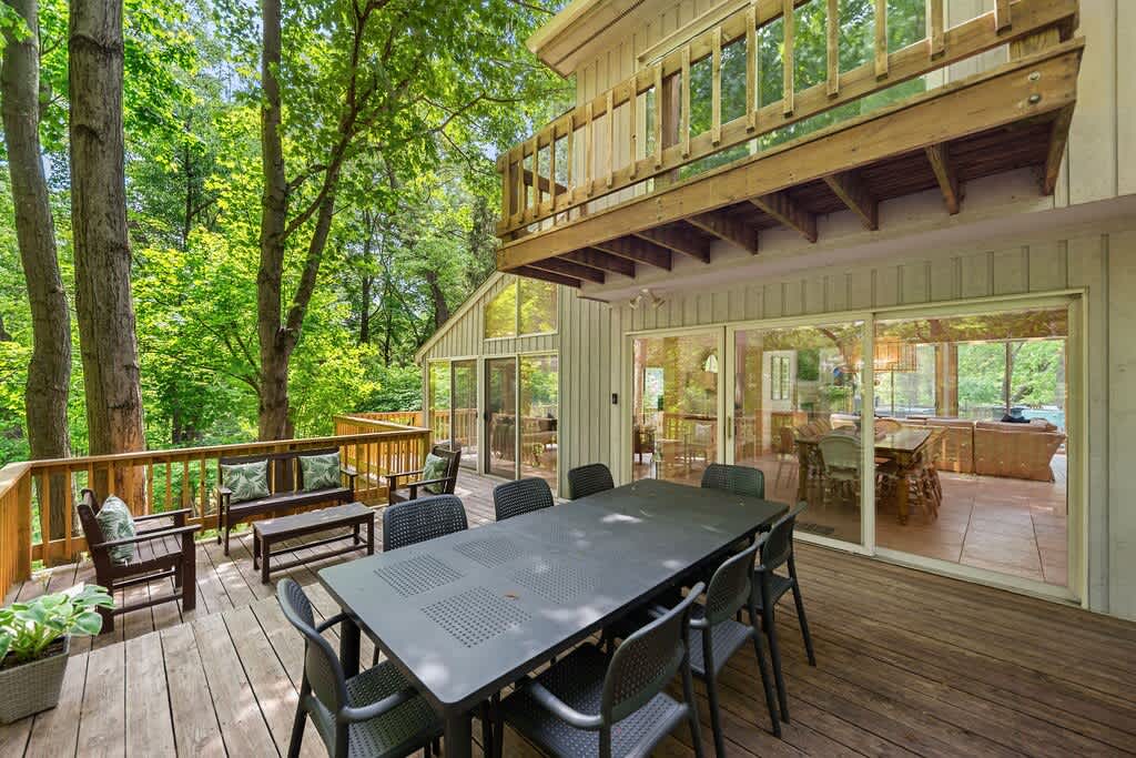 A spacious wooden deck features a large black outdoor dining table with matching chairs, surrounded by lush green trees, with a glimpse into the bright interior of the house through sliding glass doors.