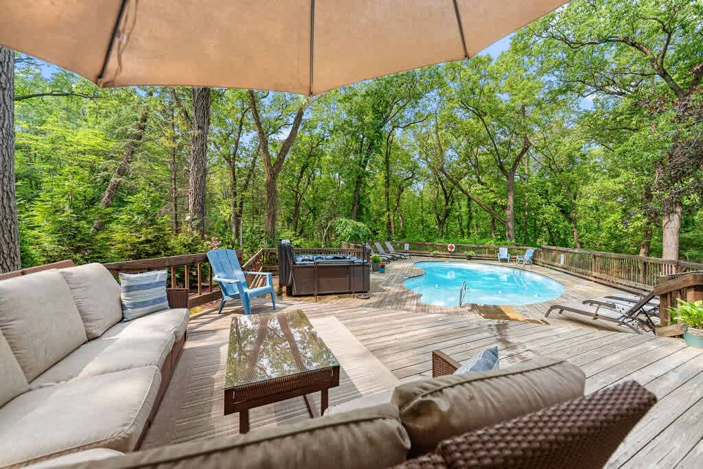 Expansive wooden deck surrounded by lush green trees, featuring an inviting outdoor seating area with a large umbrella, a hot tub, and a kidney-shaped swimming pool. Lounge chairs are scattered around the pool, creating a perfect outdoor retreat.