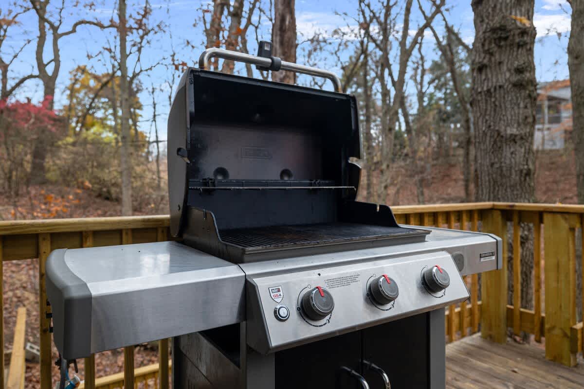 A silver and black gas grill with its lid open sits on a wooden deck, surrounded by bare trees and a blue sky in the background, ready for outdoor cooking.