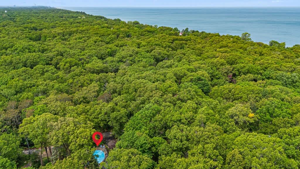 An aerial view showcases the property nestled within a dense green forest, highlighting a swimming pool and the house, with a large body of water visible in the distant background.