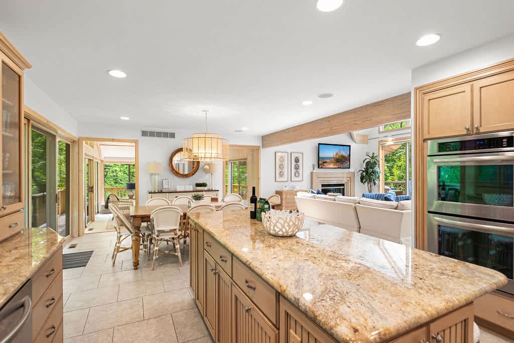 An expansive open-concept space featuring a kitchen with a large granite island and light wood cabinets, flowing into a dining area, and further into a living room with a fireplace and large windows, all connected by light tiled flooring.
