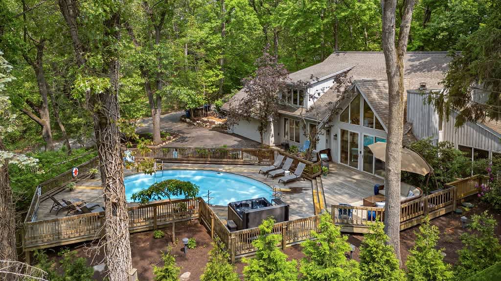 High aerial view of the modern A-frame house nestled among dense green trees, showcasing the large kidney-shaped pool and extensive wooden deck. A hot tub and various outdoor seating areas are integrated into the deck design.