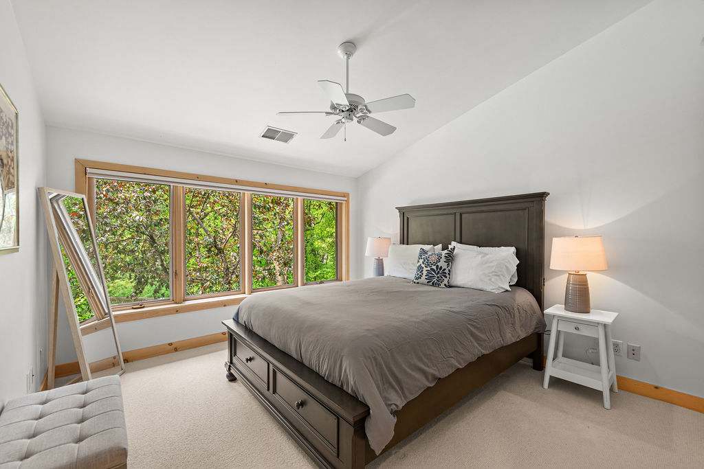 Spacious bedroom with a dark wood queen bed frame featuring storage drawers, light gray bedding, and a ceiling fan. Large windows with natural wood trim offer a lush green outdoor view, complemented by a full-length mirror and a small bench.