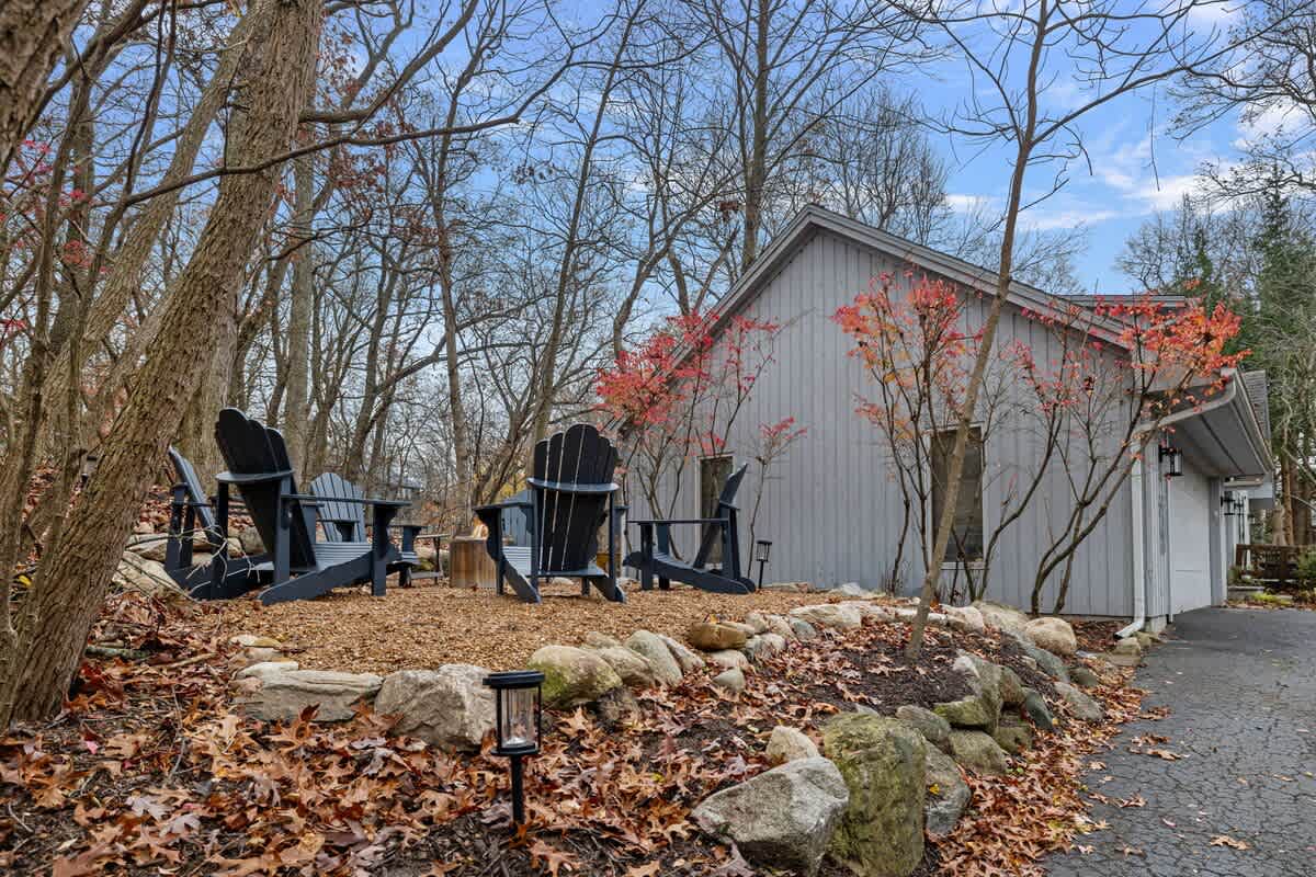 A cozy outdoor fire pit area features dark Adirondack chairs arranged around a central fire pit, set on wood chips with autumn leaves scattered around, next to a gray house.