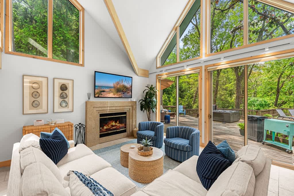 Bright living room with a vaulted ceiling and large A-frame windows offering views of lush greenery. A cream sectional sofa, two blue striped armchairs, and a stone fireplace with a mounted TV create a cozy yet open space.