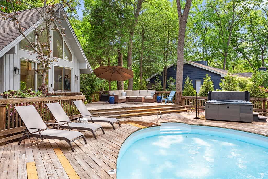 Wide shot of the property's back, highlighting a multi-level wooden deck with a kidney-shaped swimming pool and a hot tub, nestled among tall green trees. The house features a modern A-frame design with large windows and light siding.