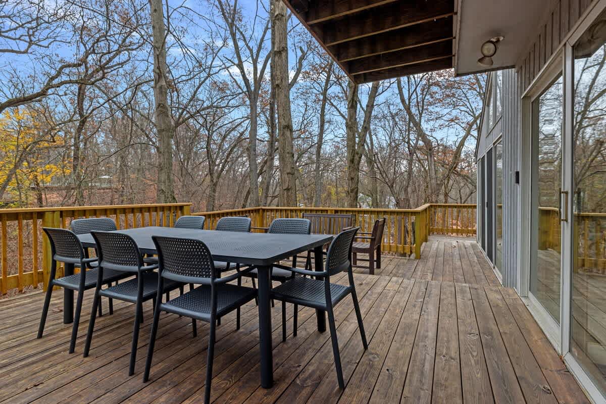 A wide wooden deck is furnished with a large black outdoor dining table and chairs, overlooking a wooded area with bare trees under a bright sky, adjacent to the house with large glass doors.