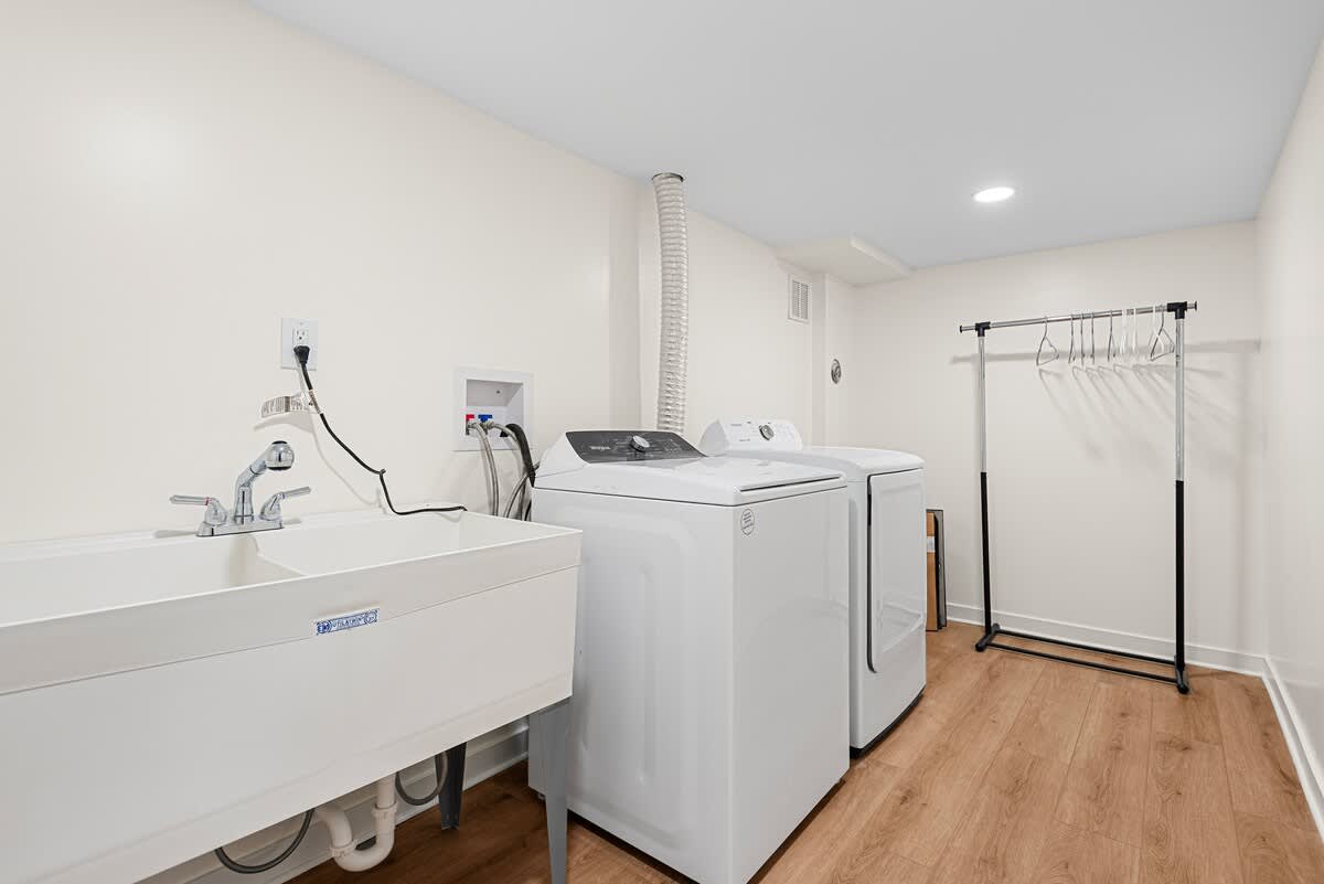 Functional laundry room equipped with a white utility sink, a modern washer and dryer, and a clothes rack. Hardwood floors and white walls create a clean workspace.