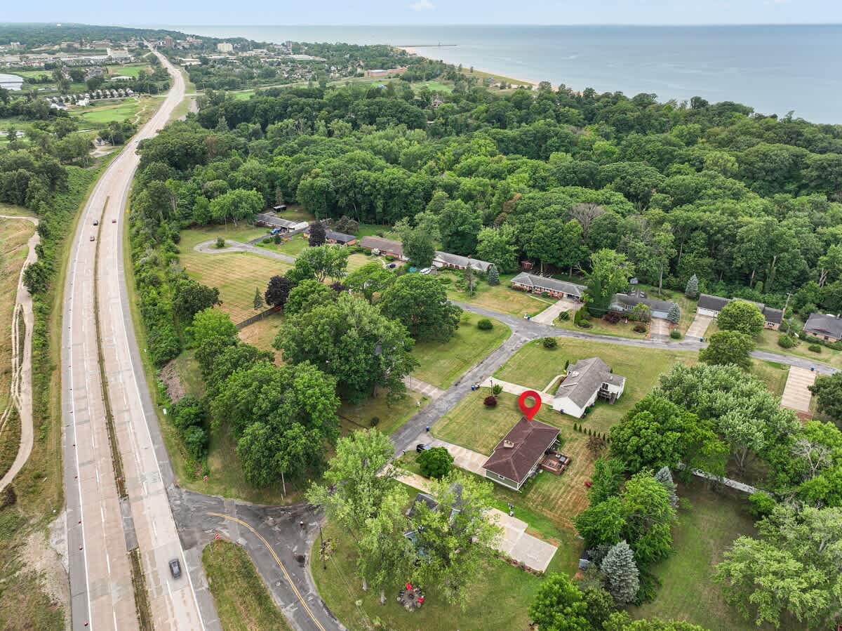 An aerial view of a residential area with a single-story house marked by a red pin, surrounded by lush green trees, with a multi-lane highway running alongside and a large body of water visible in the distance.