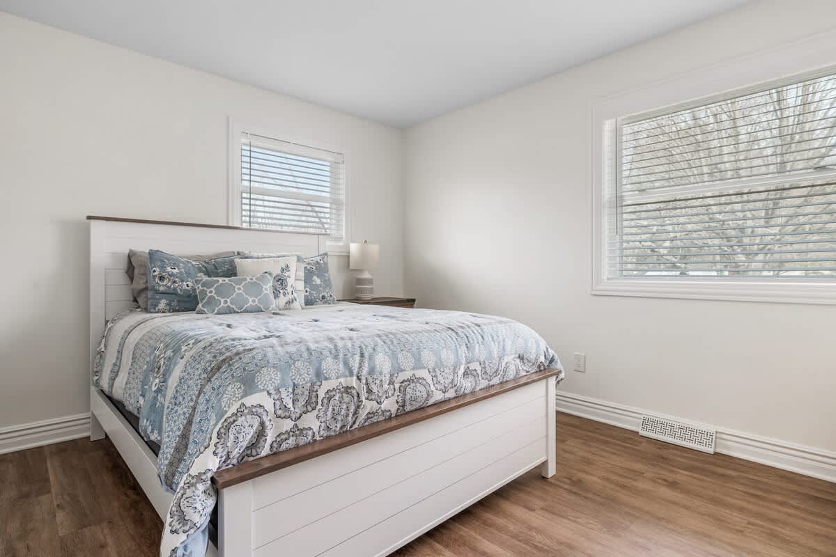 Bright bedroom with a white and wood-frame bed, blue and white bedding, and two windows with blinds, all on wood-look flooring, creating a serene atmosphere.