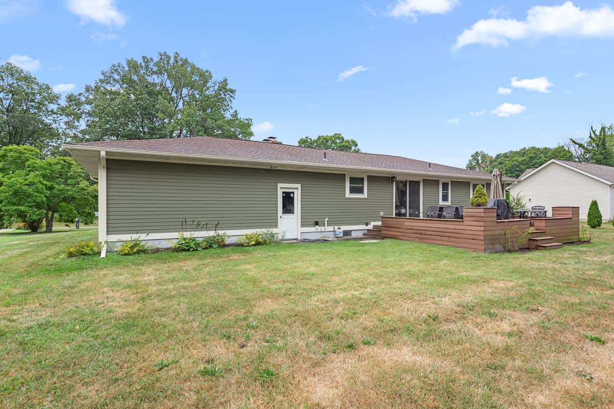 Wide shot of the back of the house, showcasing the green siding, a large wooden deck with outdoor furniture, and a well-maintained grassy yard. Mature trees frame the property.