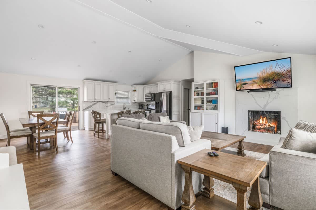 A bright, open-concept living space combines a living room with light gray sofas and a wooden coffee table, a dining area with a large wooden table, and a modern kitchen with white cabinets. Hardwood floors connect the entire area, leading to a sliding glass door.
