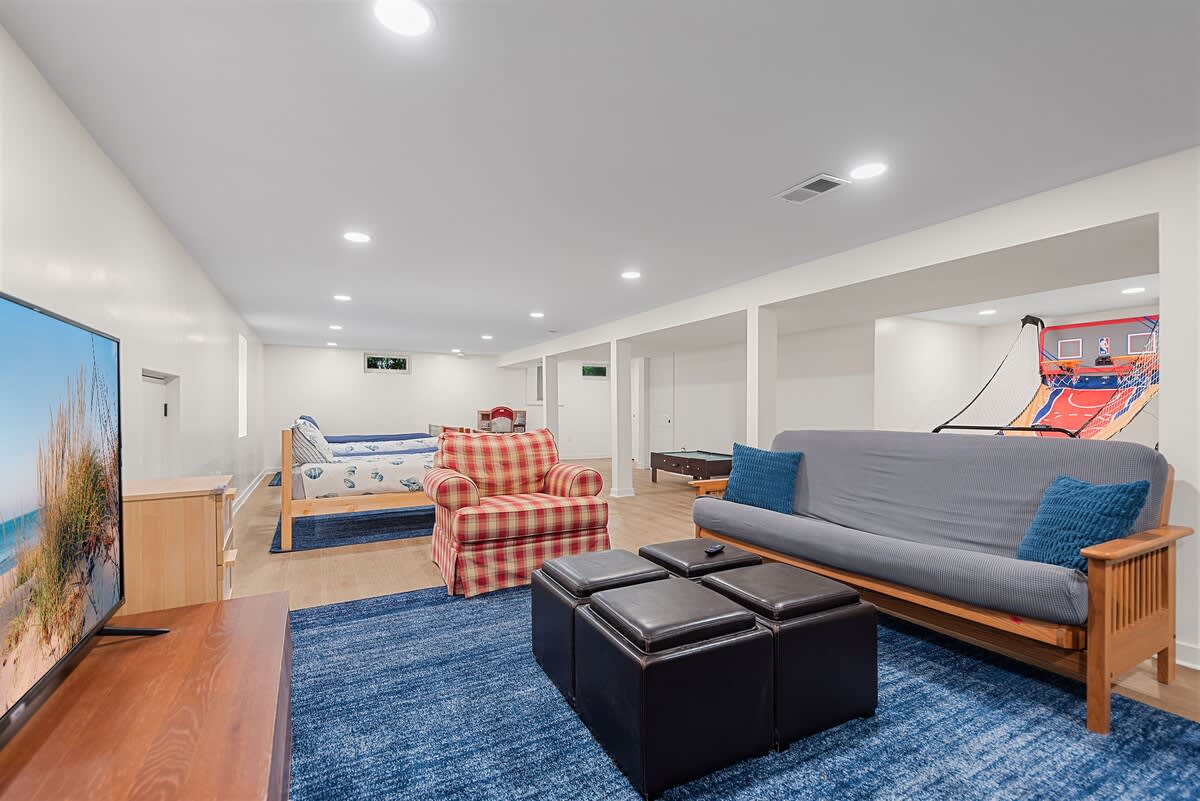 A spacious basement game room features a blue area rug, a futon, leather ottomans, and a TV on a wooden stand. In the background, there's a bunk bed, a plaid armchair, a pool table, and an arcade basketball game.