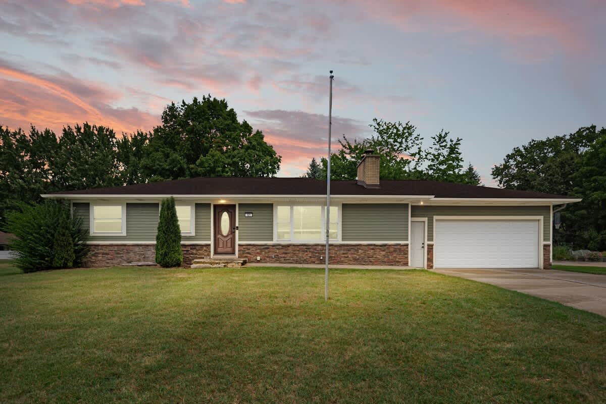 The front exterior of a single-story ranch-style house with sage green siding and stone accents, a dark brown roof, a white garage door, and a well-maintained green lawn under a dramatic sunset sky.