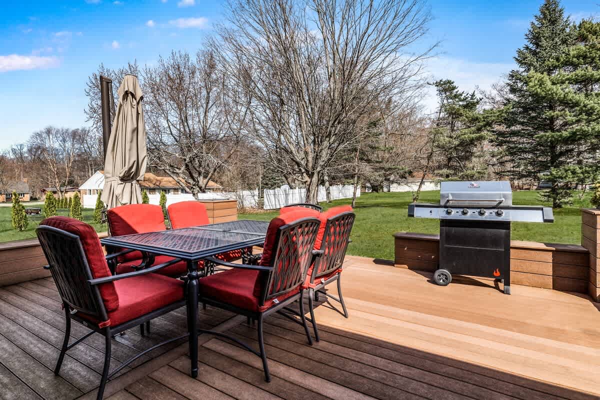 A spacious wooden deck features a black metal dining table with red cushioned chairs and a large BBQ grill. The deck overlooks a grassy backyard with mature trees and a white fence in the distance.