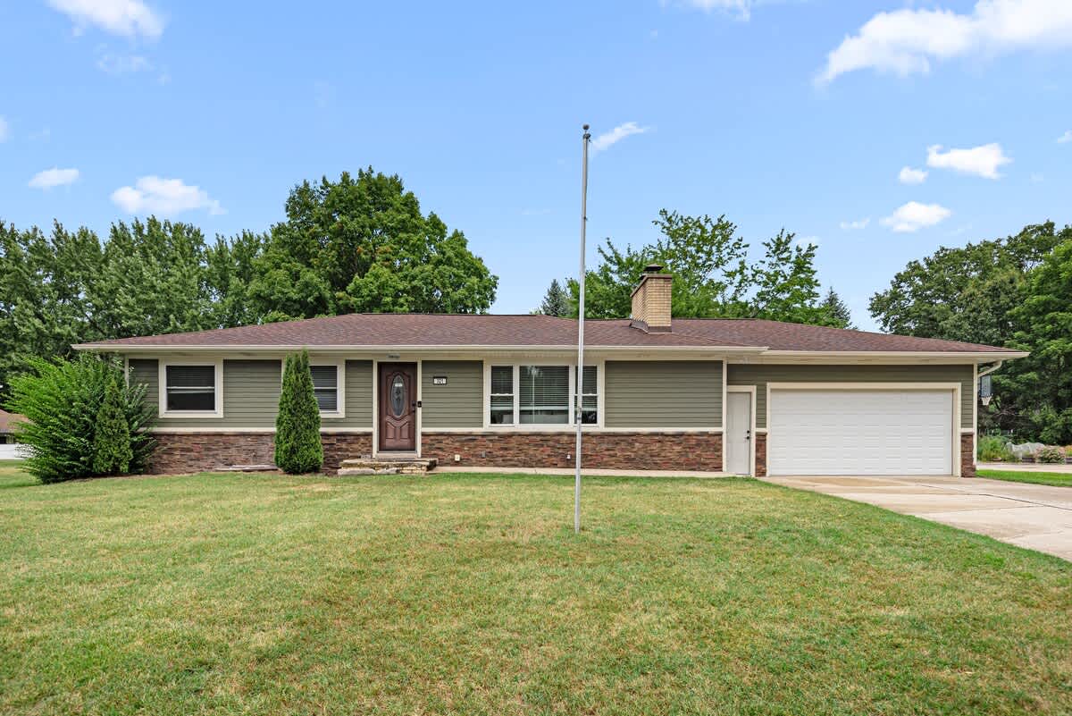 The front exterior of a single-story house features olive green siding, a brown stone facade, and a white garage door. A well-maintained green lawn and mature shrubs are visible under a bright blue sky.