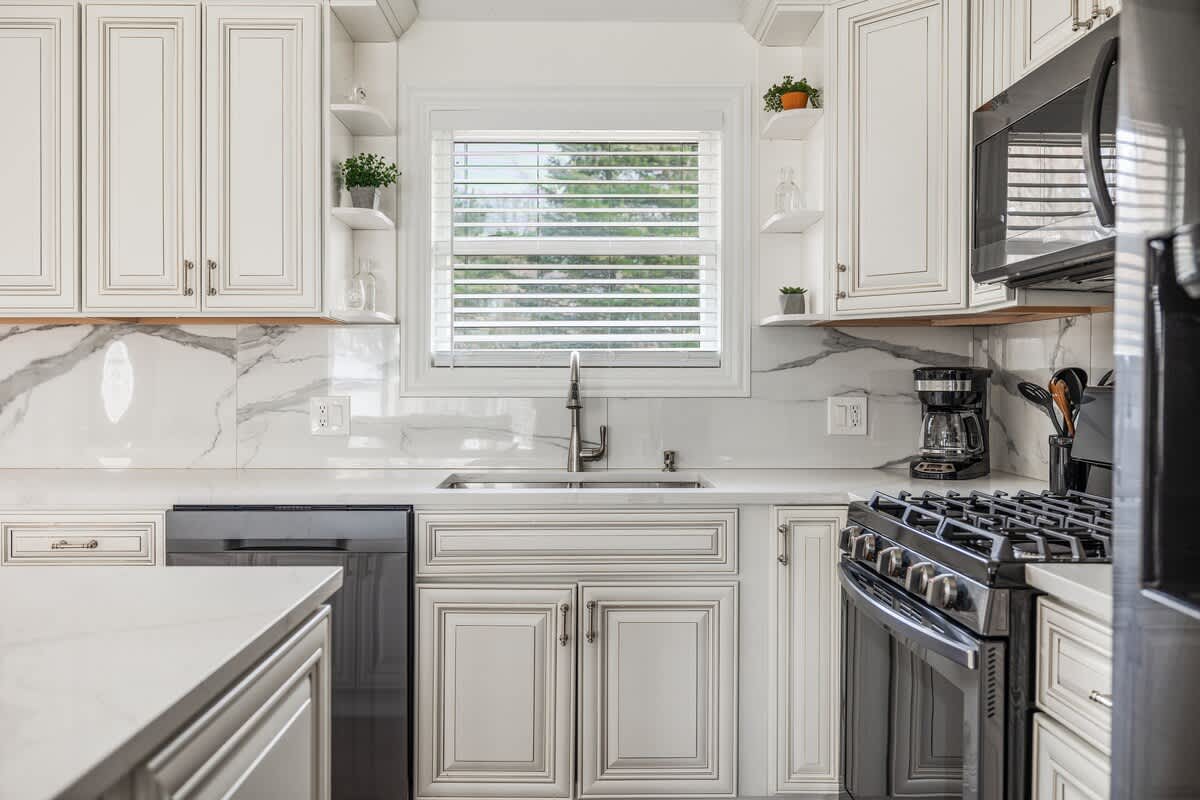 Close-up of a modern kitchen featuring white shaker cabinets, white marble-patterned backsplash, a stainless steel sink, and black appliances including a dishwasher and stove.