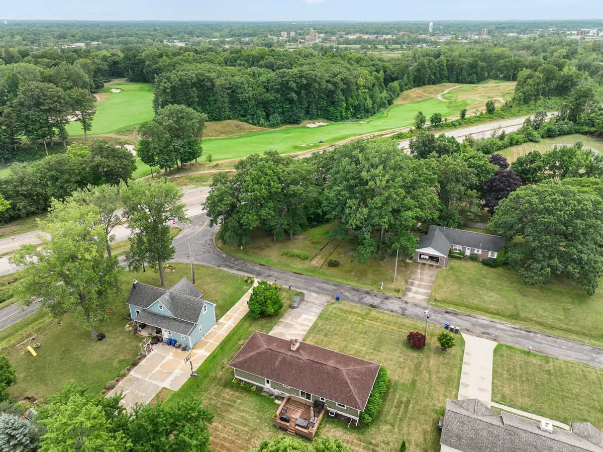 An aerial view showcasing a residential neighborhood nestled among dense green trees, with a manicured golf course visible in the background and a highway running through the landscape.
