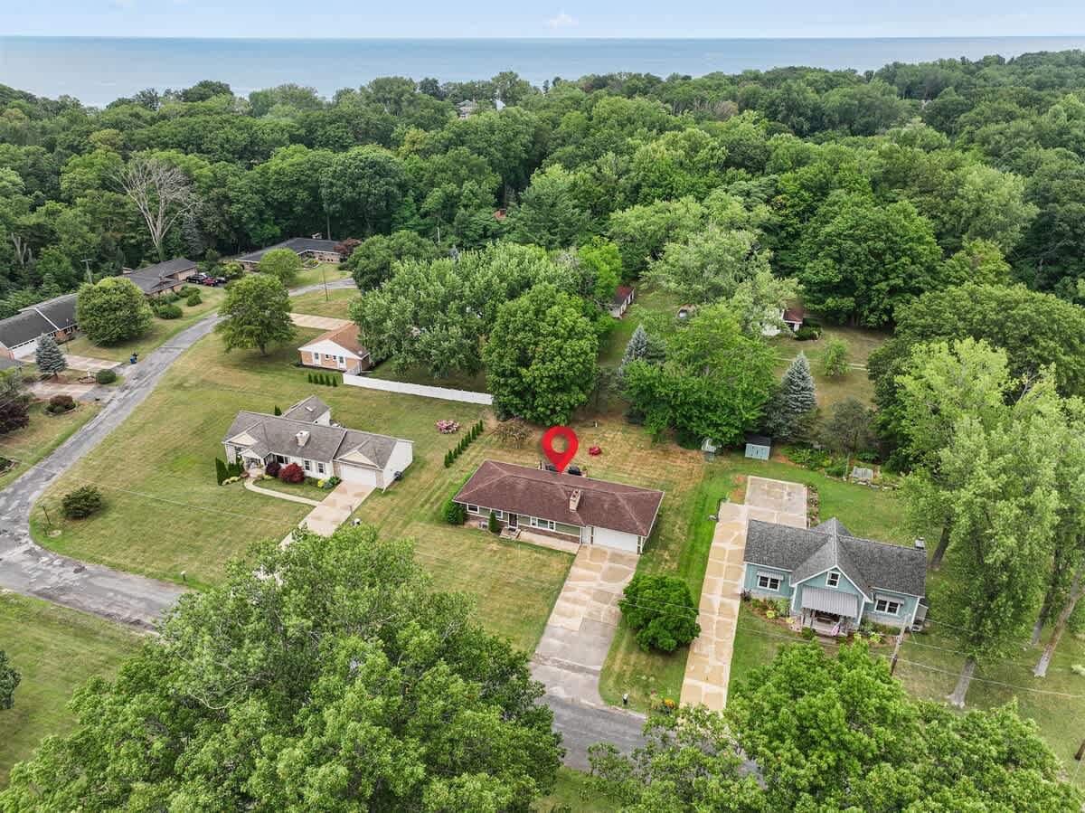 An aerial perspective of a single-story house marked with a red pin, situated in a green, tree-filled neighborhood with other homes, and a large body of water visible on the distant horizon.
