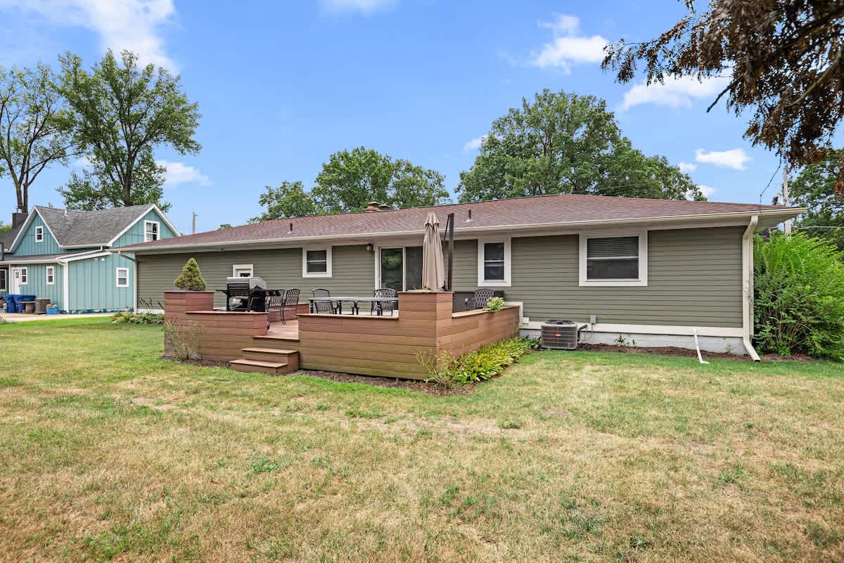 Rear view of a single-story house with green siding and a large wooden deck furnished with outdoor seating and a grill. The deck overlooks a spacious grassy backyard under a blue sky.