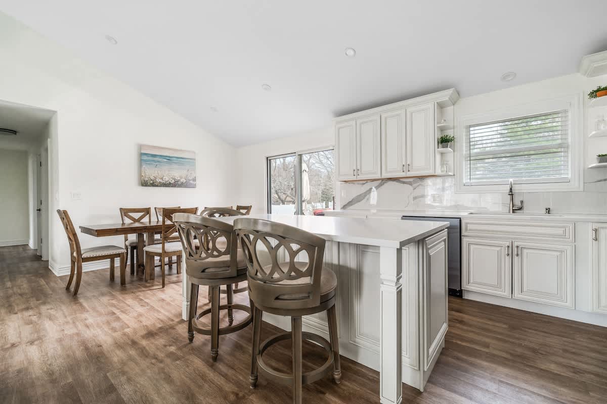 Open concept kitchen and dining area with white shaker cabinets, a large island with seating, and a wooden dining table, all on dark wood-look flooring under a vaulted ceiling.