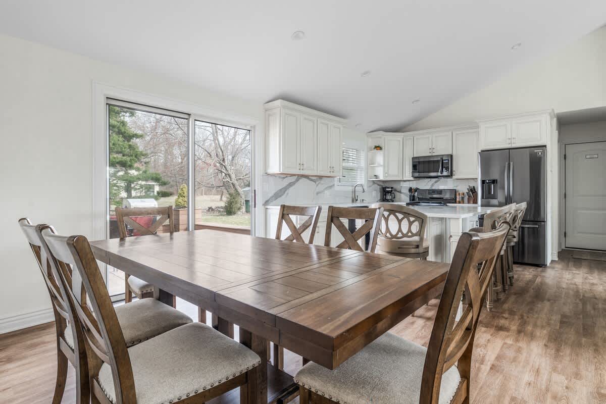 A spacious dining area features a large wooden dining table with six upholstered chairs, set against a backdrop of a modern kitchen with white cabinets. A sliding glass door leads to an outdoor deck, providing ample natural light.
