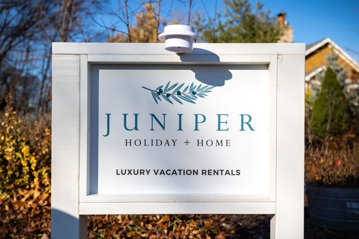 A white wooden sign with 'JUNIPER HOLIDAY + HOME' and 'LUXURY VACATION RENTALS' printed in blue and black text, featuring a juniper branch logo, set outdoors with blurred green and brown foliage in the background.