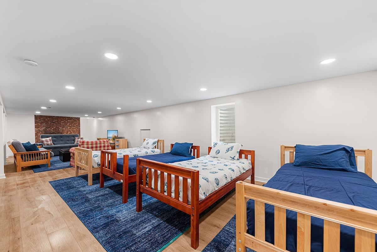 A spacious basement recreation room with light wood-look flooring and white walls, featuring three wooden twin beds with blue and white patterned bedding, and a cozy seating area with a gray sofa and wooden chairs in the background.