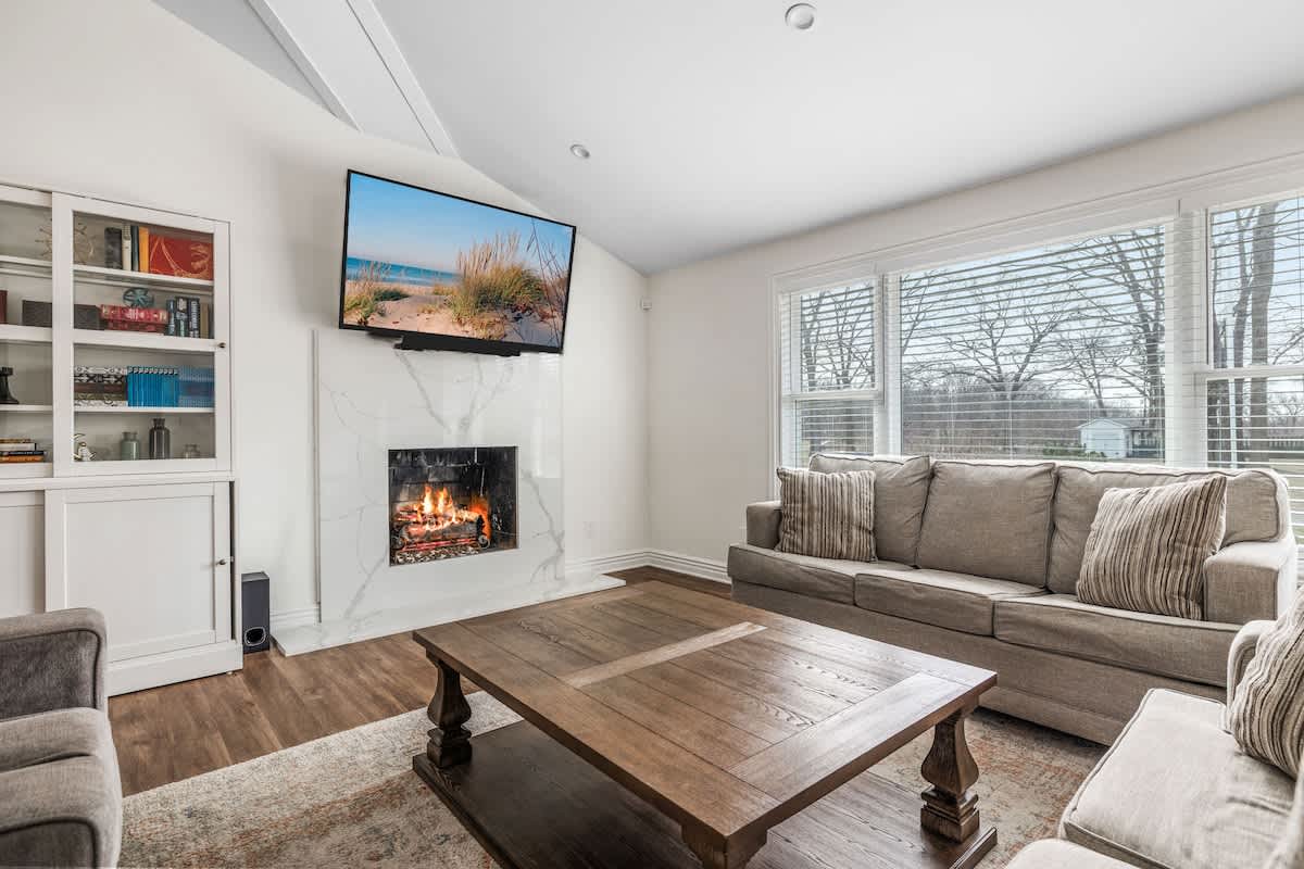 A cozy living room features a light gray sofa, a large wooden coffee table, and a white marble-look fireplace with a wall-mounted TV. Large windows with blinds offer natural light and a view of the bare trees outside.