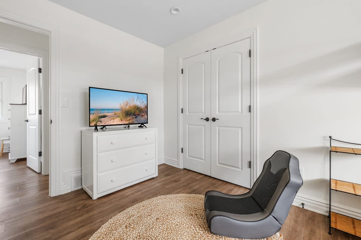 View of the kids' bedroom showing a white dresser with a TV, a gray gaming chair on a jute rug, and double closet doors. Hardwood floors and white walls maintain a bright feel.