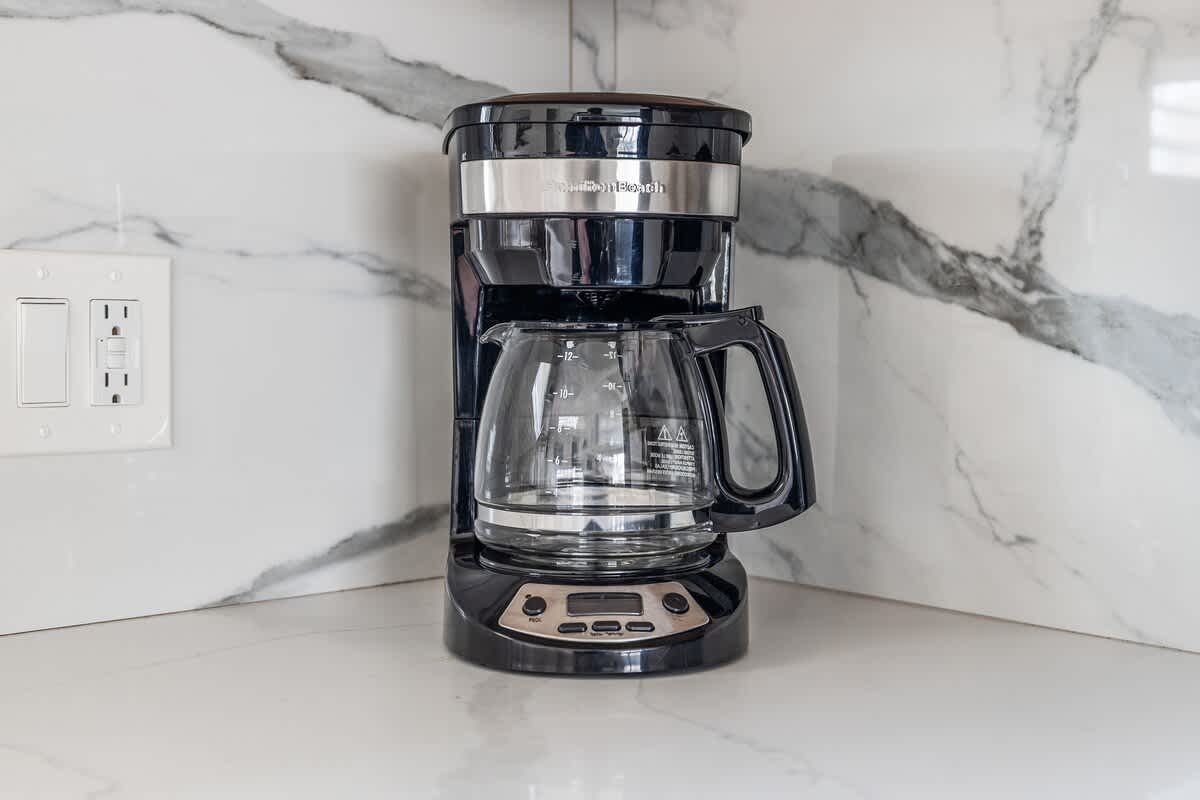 Close-up of a black coffee maker on a white countertop with a white marble-patterned backsplash, next to a white electrical outlet.