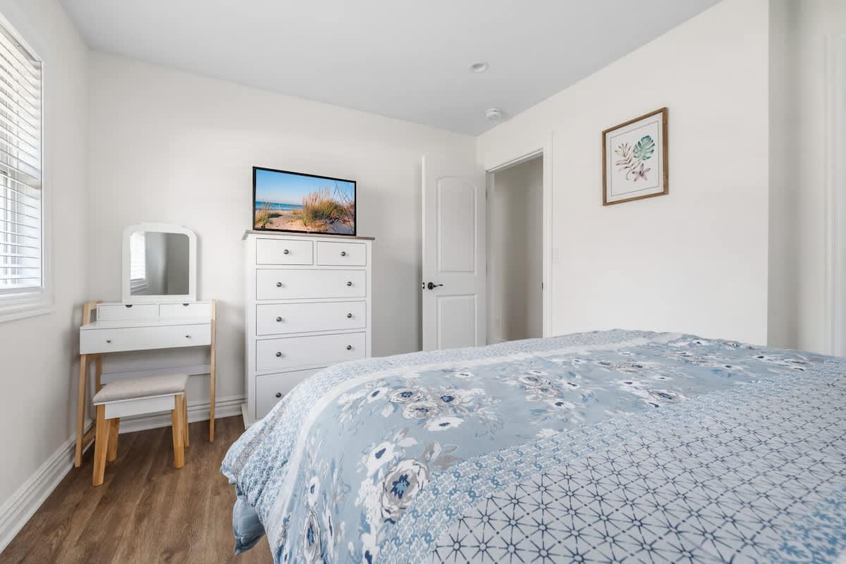 Bright bedroom with white walls, a bed with a blue floral duvet, a white dresser with a TV, and a modern white vanity with a mirror. Hardwood floors add warmth to the clean aesthetic.